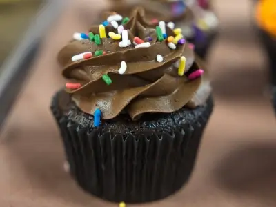 Chocolate cupcake with swirled chocolate frosting, topped with colorful sprinkles, sitting on a brown surface.