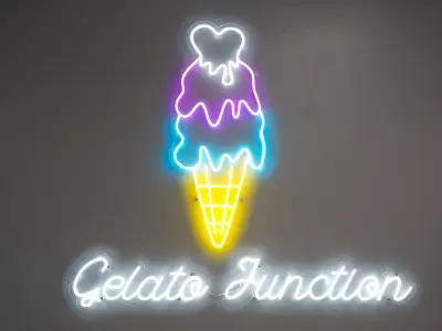 Neon sign of an ice cream cone with three scoops in pink, purple, and blue, above the words “Gelato Junction” in white cursive on a gray background.