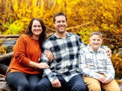 A woman, man, and boy sit closely together outdoors in front of vibrant autumn foliage, all smiling at the camera.
